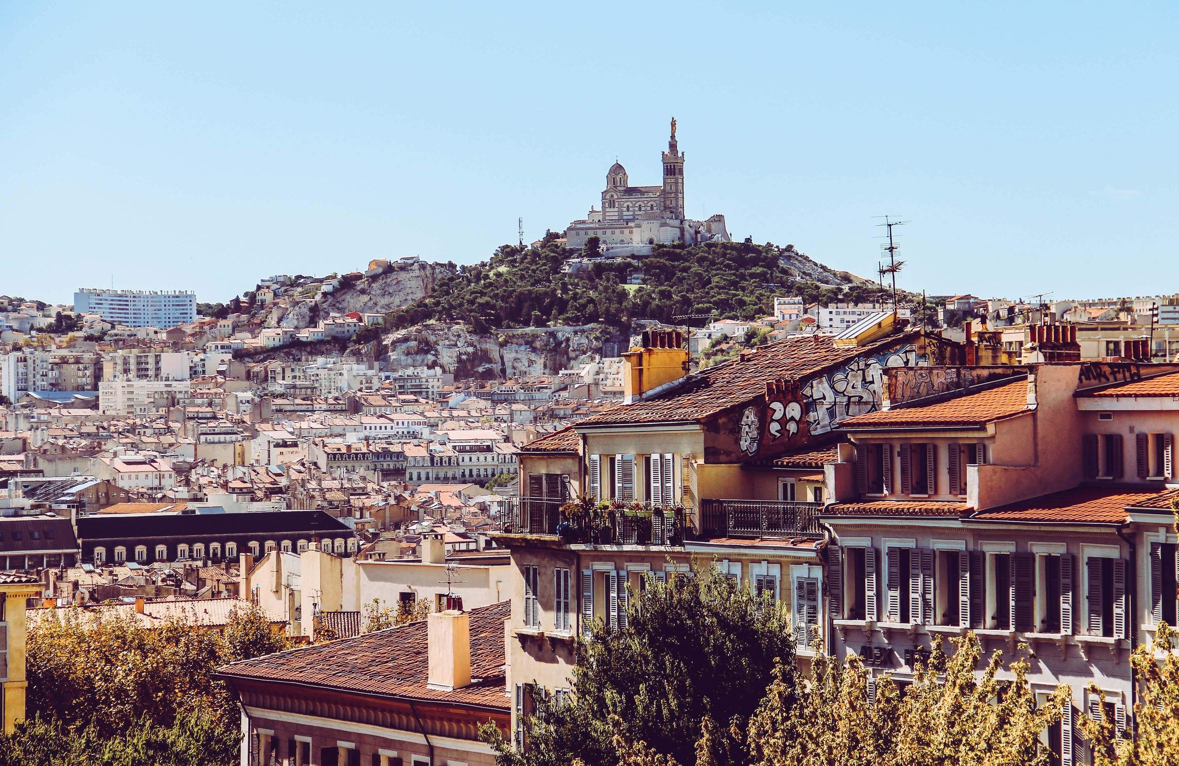 vue sur notre Dame de la Garde avec en arrière plan une coline surmonté d'une église et en premier plan la vue sur la ville