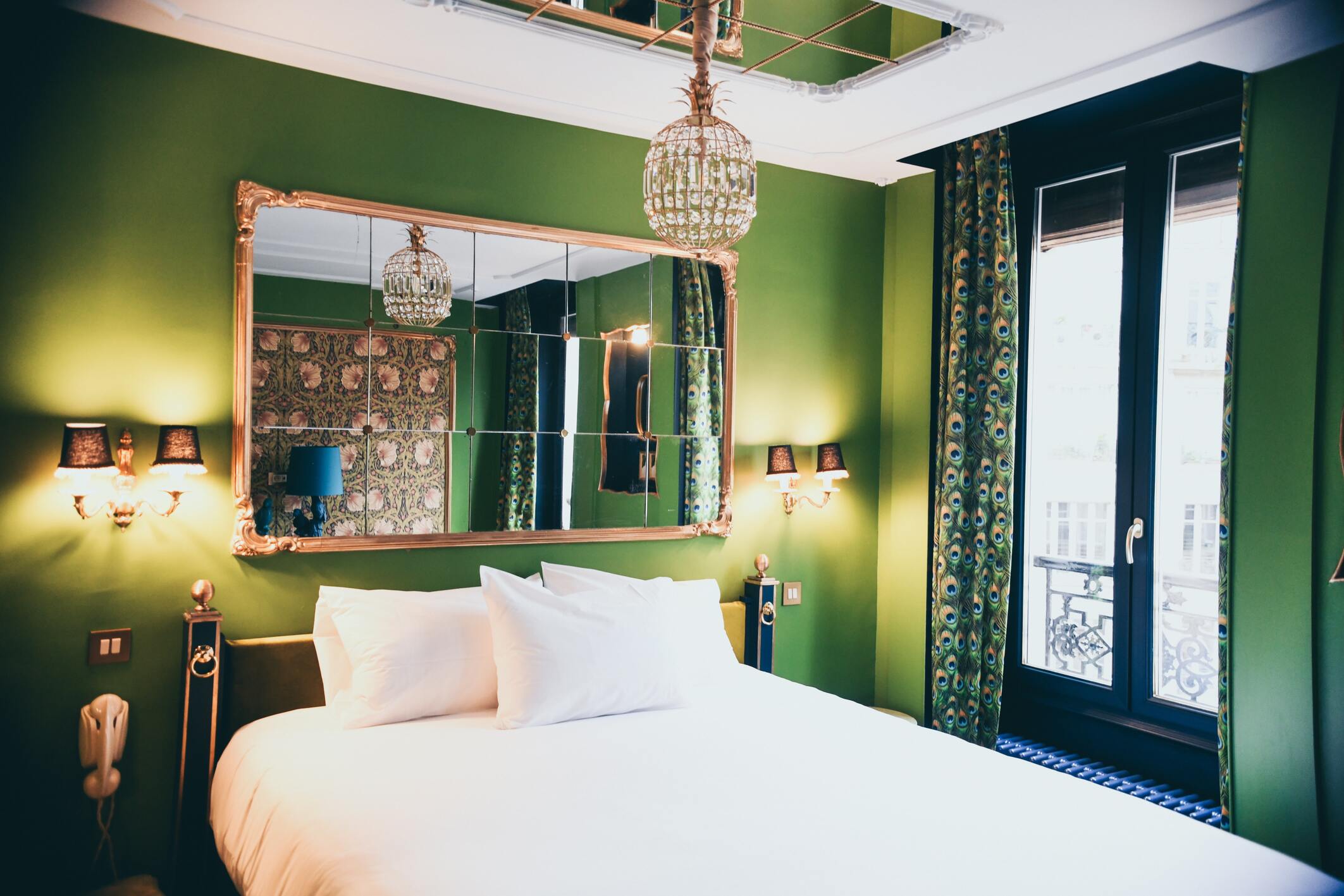 chambre verte kaki avec un lit double, un grand miroir au dessus du lit et une porte fenêtre avec un petit balcon 